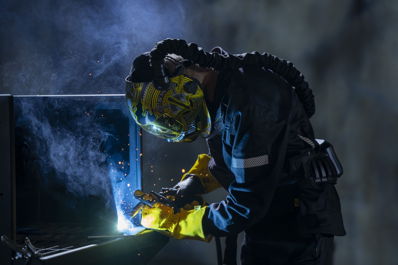 In a dark room, a welder uses a Rustler MIG PRO compact 350C pulse welder on a machine, sparks flying around him.