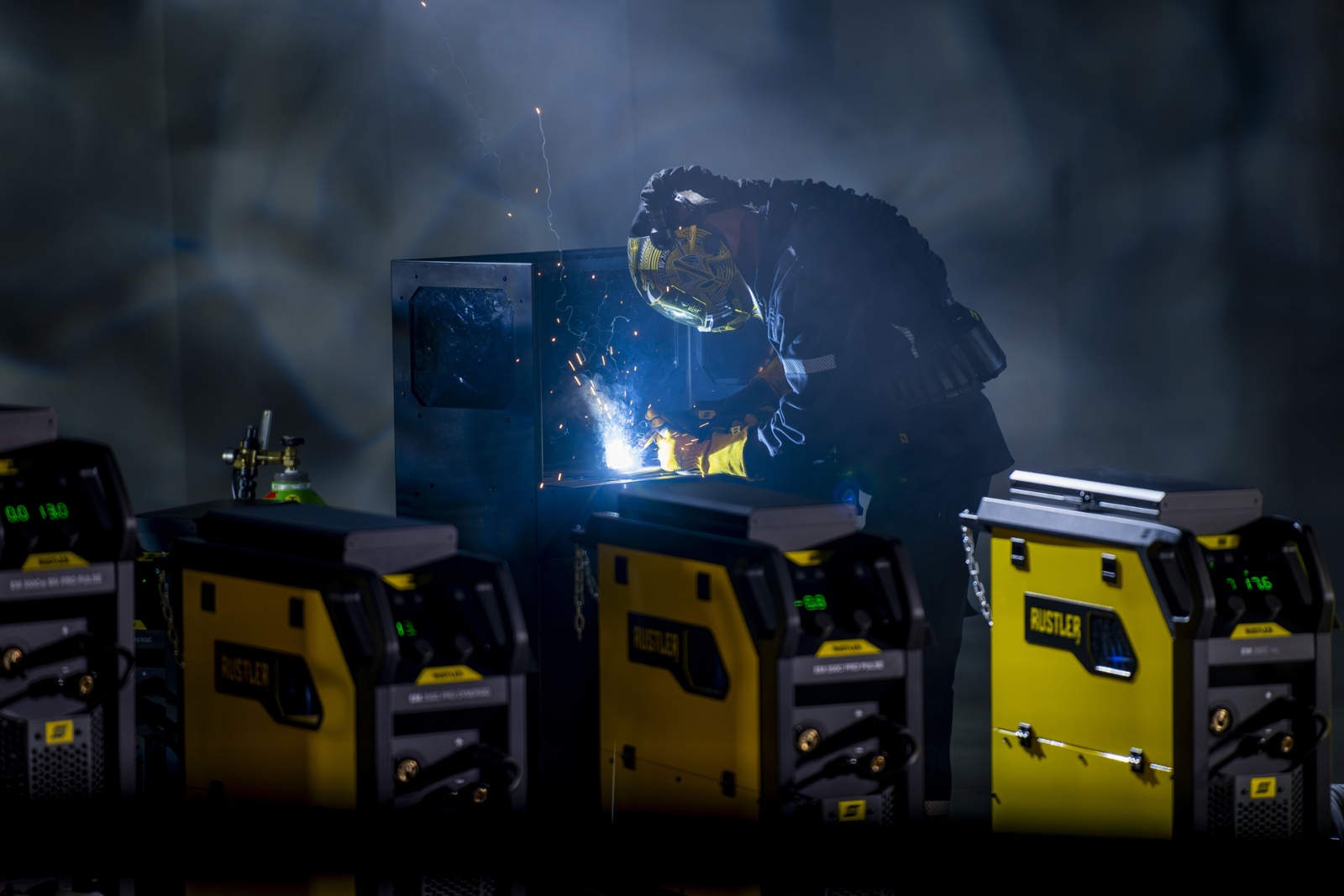A man operates a Rustler MIG PRO compact 350C welder in a shadowy room, concentrating on the welding process.