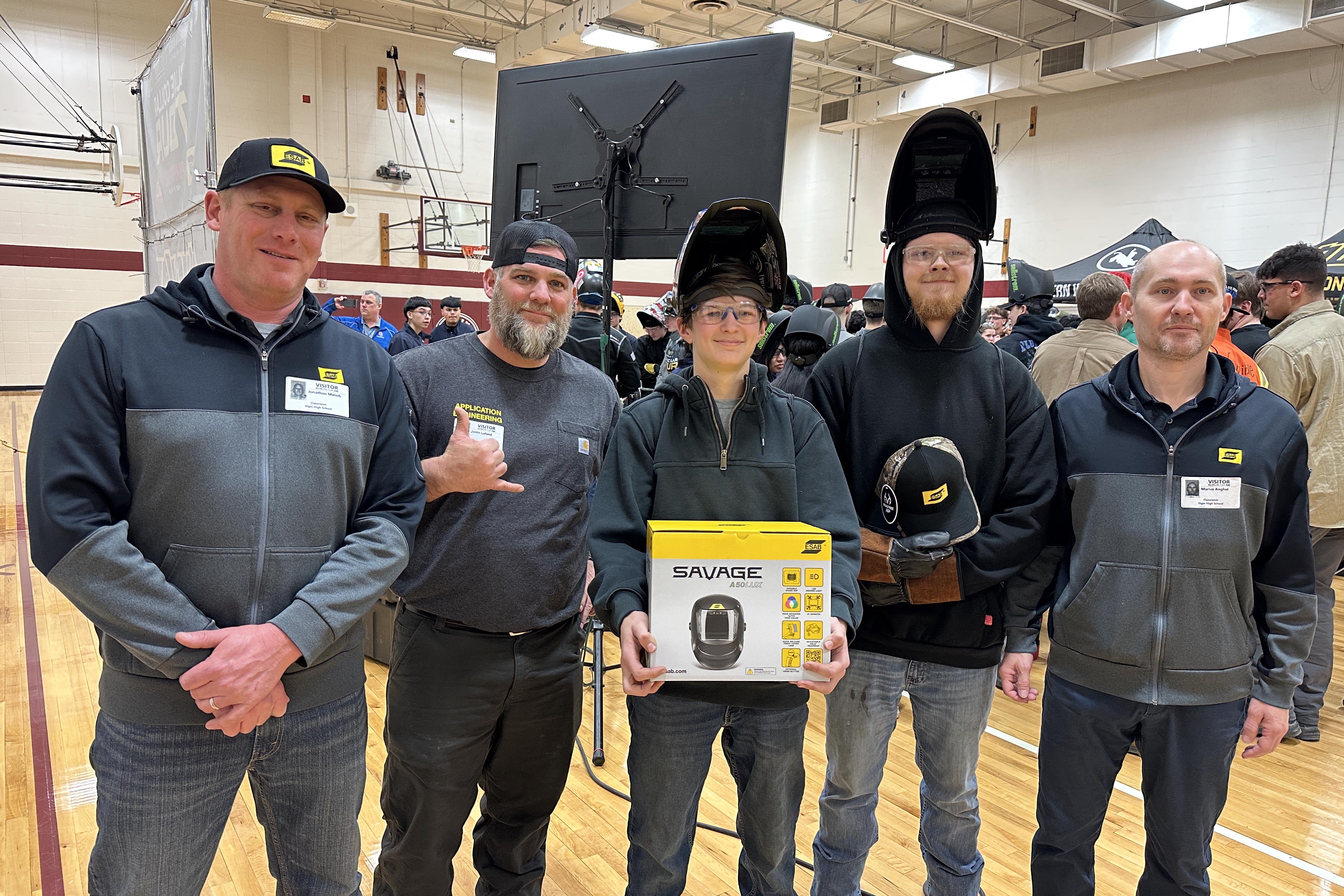 Five people stands posing for a photo. high school junior Lou Jensen in the center holds a Savage welding helmet box.